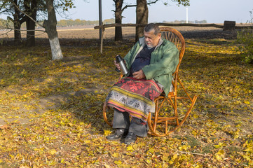 Senior man sitting in wicker chair and ready to drick tea at sunny autumnal day.