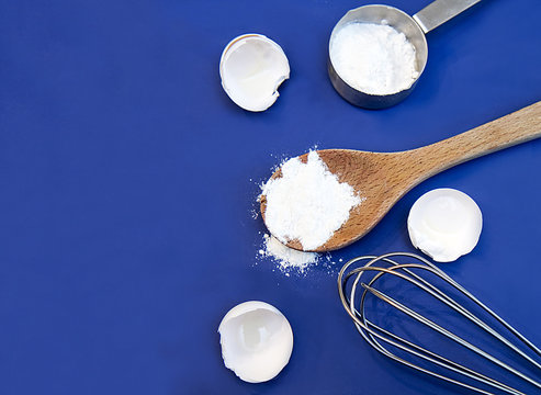 Blue background for cooking or baking using ingredients flour and eggs.  Imange consists of egg shells, cooking tools as whisk, measuring cup and wooden spoon holding flour.