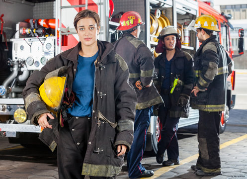 Confident Firewoman Holding Helmet While Colleagues Discussing B