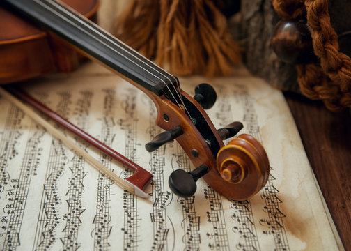 Old wooden string instrument (violin) resting on classical sheet music. Focus is on the scroll, tuning pegs, bow, and part of the neck.
