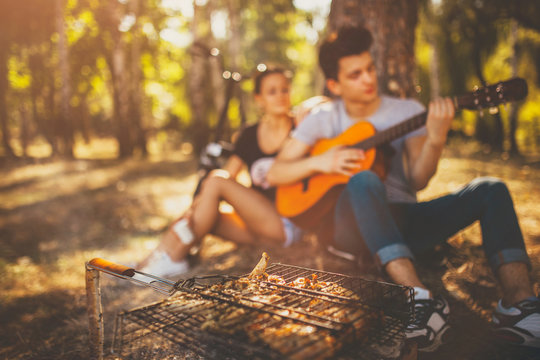 Picnic. Grilling meat and playing guitar. Defocused background. Teenagers loving boy and girl sitting together by a tree on weekend sunny day.