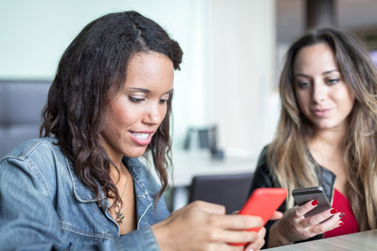 Two young women friends are on the table in a bar, both are using their mobile phone, the blonde girl looks at the phone in the hands of her friend