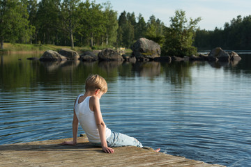 Fototapeta premium Boy relaxing on a jetty. He's sitting in the sunshine and looking out over the beautiful lake Skiren in the swedish countryside.