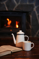 Hot tea or coffee in mug, book and candles on vintage wood table. Fireplace as background