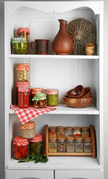 Jars with pickled vegetables and beans, spices, book of recipes and kitchen utensils on shelves of cupboard