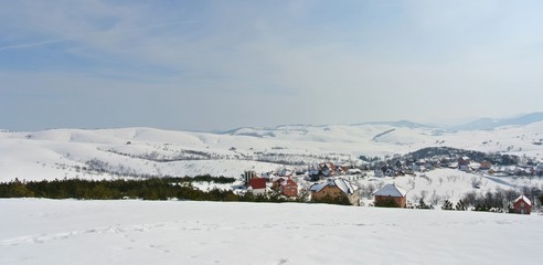 Panoramic image of beautiful, idyllic snowy winter landscape in the mountains, on a crisp sunny morning, with snow-covered rooftops of a small picturesque village.