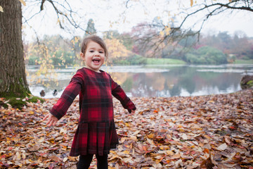 A toddler girl in red tartan dress playing with falling leaves and sticks standing in front of the lake holding her hair