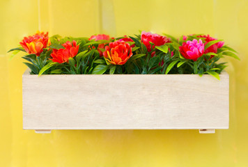 Artificial flowers in wooden box hang on the bright yellow wall.