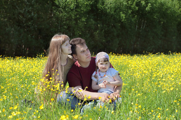 Happy woman, man and daughter sit among yellow flowers at meadow