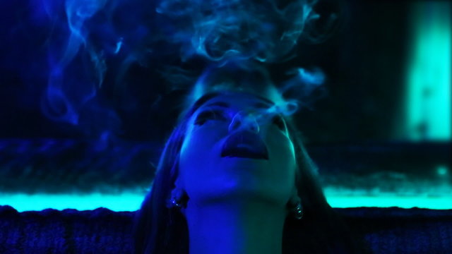 Club Party – Nightlife. Beautiful Sexy Young Glamorous Woman Relaxing in the Chill-out in Nightclub Exhales Smoke. Chilling/Relaxation Stylish concept. Home Party. Blue Mystic illuminated