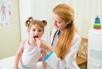 Pediatrician woman giving medicament to little girl with a spoon. Kid is opening mouth. Hospital room on the background.