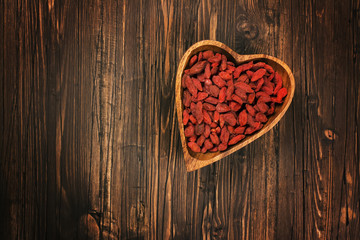 Goji berries in a heart-shaped wooden bowl over wooden background. Top view. Space for text. Toned image