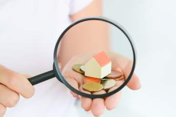 Real estate concept - coins and house architectural model in woman hand under magnifying glass