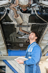Fototapeta na wymiar Mechanic With Clipboard Examining Under Car In Garage