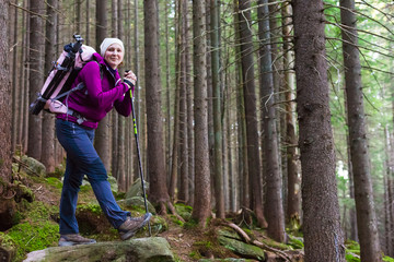 Naklejka na ściany i meble Female Hiker Staying inside Deep Old Forest on Moss Stone with Backpack and Trekking Pole Smiling and Joyful