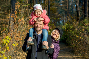 Naklejka na ściany i meble Happy Family on the Walk in Autumnal Forest Father Mother Little Baby Daughter Together on Weekend Walk on Autumnal Colors Forest Sunny Day Smiling Faces Child Sitting on Daddy Shoulders