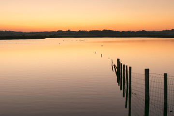 Fototapeta na wymiar Orange and yellow sunset over still water lake with wooden fence posts to right
