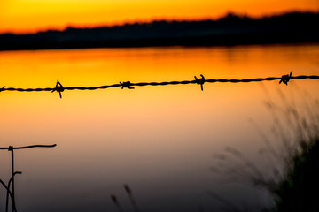 Barbed wire in front of orange and mauve sunset over still water lake