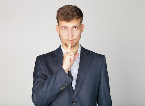 Young business man in suit stands confidently and shows the sign of silence by his finger near mouth.