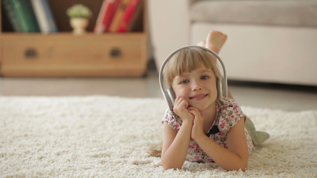 Cheerful little girl lying on carpet in living room listening to music on headphones and smiling at camera