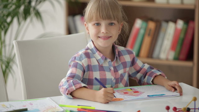 Cute little girl sitting at table drawing with colored pencils and showing her painting at camera