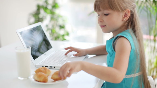 Cute little girl sitting at table eating cakes and using laptop