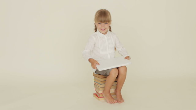 Pretty little girl sitting on pile of books holding laptop closing it and showing thumb up at camera