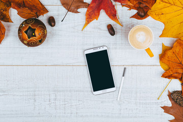 Colorful background image with fallen autumn leaves, coffee cup and mobile phone on white wooden table.