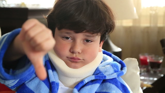 Portrait of a painful little boy with a neck brace looking at the camera and showing thumbs down