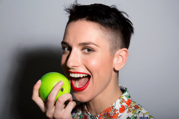 Brunette with short hair biting a green apple on a light background.