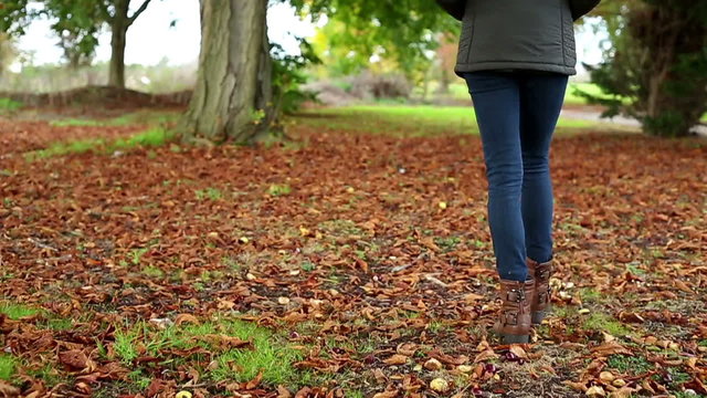 Young woman walking through dried leaves on a autumn day in slow motion kicking up the leaves