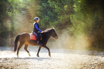 A young girl riding her pony during riding lesson, outside. Natural sun rays shining in dust during...