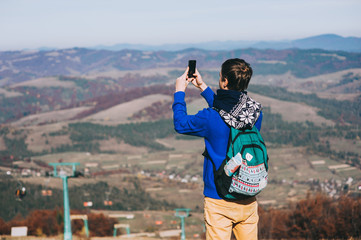 Fototapeta na wymiar Young hipster taking photo by smart-phone on the peak of mountai