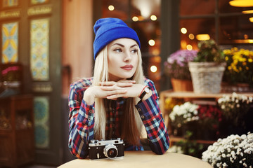 Obraz premium Portrait of beautiful stylish young woman sitting in street cafe and drinking coffee. Hipster with old retro camera. Model looking aside. Lights and flowers background. City lifestyle. Toned