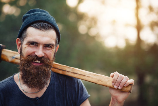 Lumberjack brutal bearded man in warm hat with a cigarette and an ax in the autumn forest at sunset
