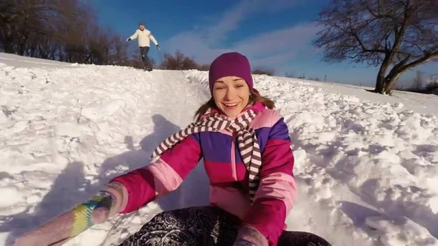 Cute young adult riding down a steep hill on a sledge. Boyfriend waving hand and smiling