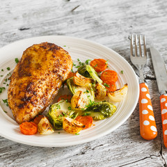 baked chicken breast with brussels sprouts, onions and carrots on a white plate on wooden surface. Healthy food