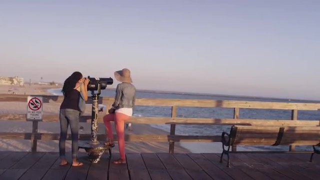 Black women best friends looking through telescope on pier 