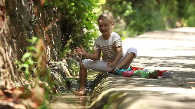 Girl sitting beside a stream and launching a lot of paper boats on water