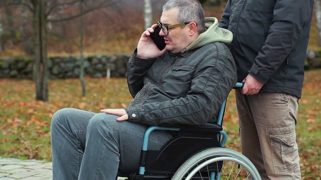 Disabled man talking on smartphone in wheelchair with assistant behind