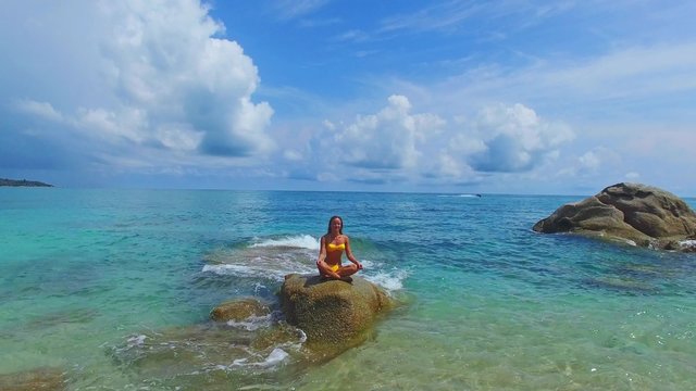 Young beautiful yoga teacher in yellow bikini doing yoga on the rock in the sea. Clear water on Samui island in Thailand. Aerial veiw from the drone.