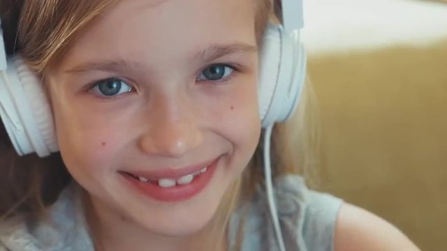 Portrait blonde girl 8 years listening music in headphones and looking and smiling at camera. Zooming