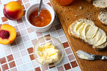 Tasty jam in the jar and bowl, ripe peaches, bread with butter and wooden tablet on mosaic background