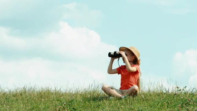 Young naturalist through binoculars watching wildlife and looking at camera. Girl sitting on the grass against the sky