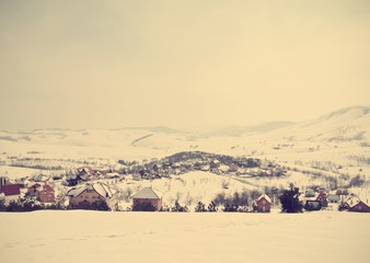 Idyllic rural snowy winter landscape in a mountain village, with snow-covered rooftops. Image filtered in faded, retro, Instagram style; nostalgic vintage rural winter concept.