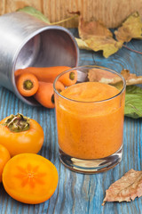 Glass of persimmon smoothie with carrot juice flavored cinnamon, decorated by ingredients and fallen leaves on blue wooden board
