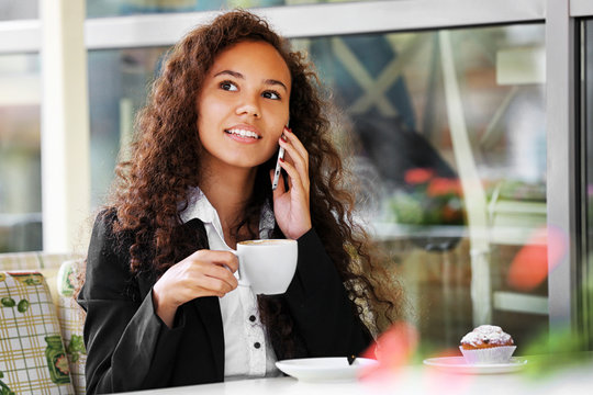 Young smiling woman speaking by cellphone and drinking coffee at the restaurant's terrace