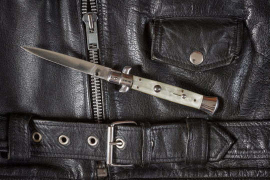 Opened flick knife on a black leather jacket with zip, belt and pocket