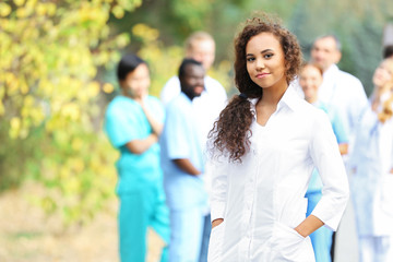 Obraz premium Attractive young woman doctor with clipboard in hands against group of medics, outdoors