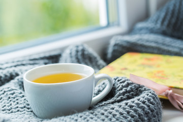 White cup of chamomile tea with grey scarf on the windowsill.